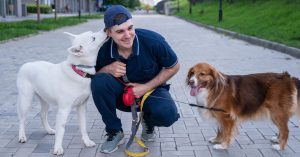 A man crouches down on a sidewalk while holding two leashes. One dog licks his ear and the other stands still while panting.