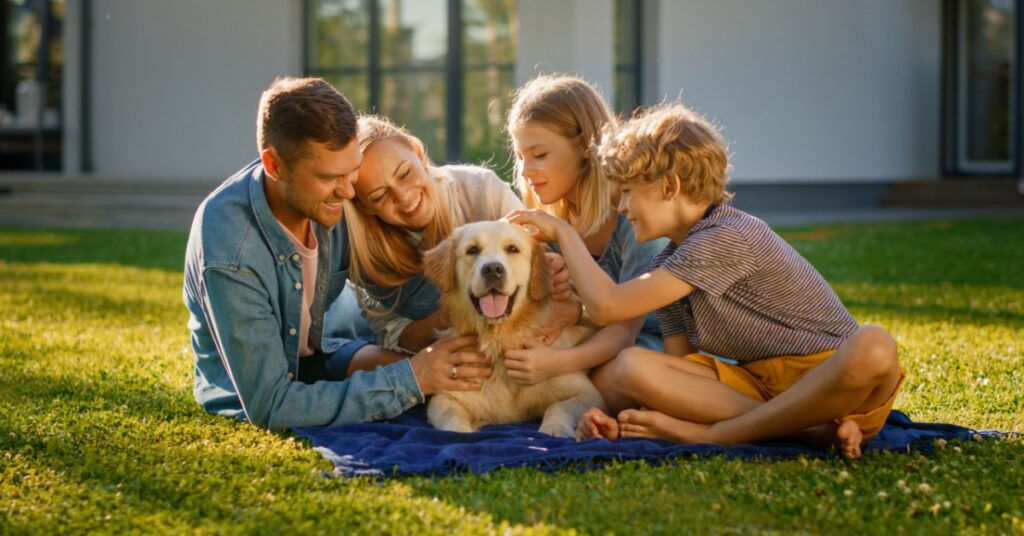 Two parents lie down beside their young kids and golden retriever dog. They're all on top of a blanket on a grassy lawn.