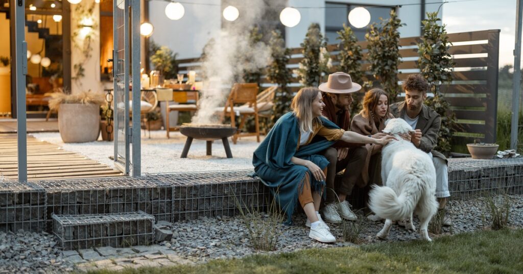 Four adults are sitting on a patio beside a fire pit and string lights while petting a large white dog.